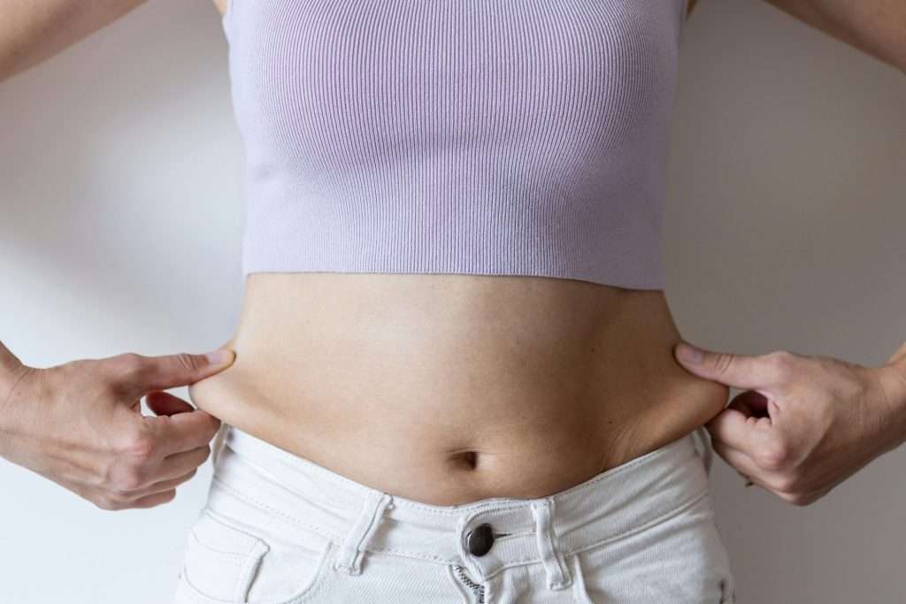 Cropped image of overweight woman, tucking, hiding fat naked big excessive belly with navel in white